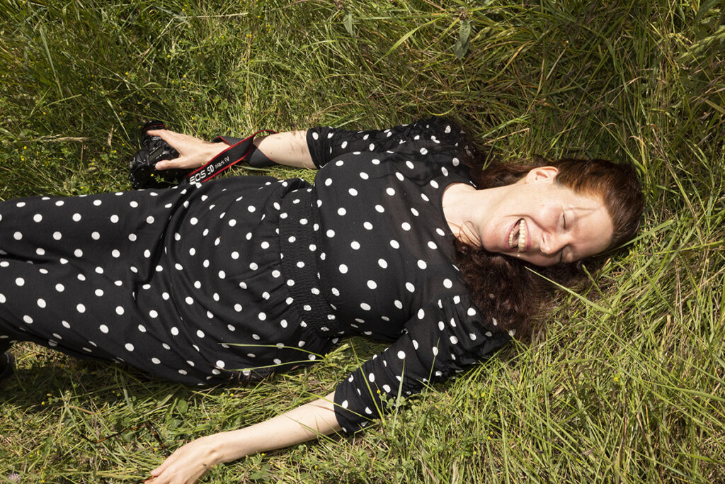 Une femme habillée d'une robe noire à pois blanc, couchée dans l'herbe haute, avec un appareil photo dans sa main droite, rit.
