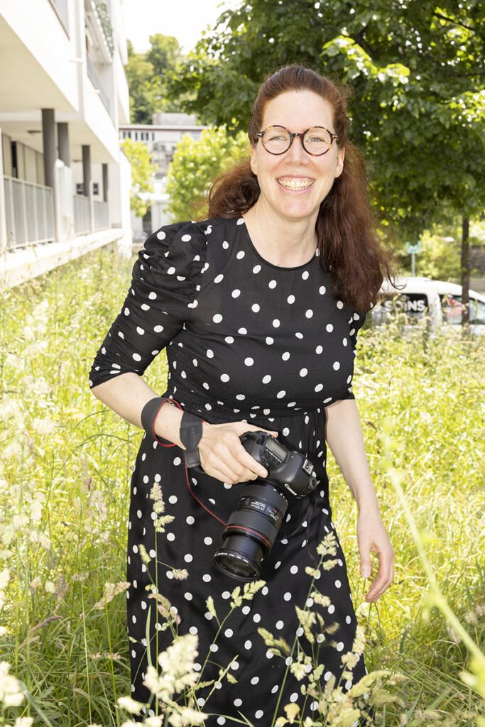 Une femme habillée d'une robe noire à pois blanc, dans l'herbe haute, avec un appareil photo dans sa main droite, sourit.
